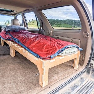May include: A wooden platform bed frame with a red sleeping bag and a gray pillow inside a vehicle. The sleeping bag has blue trim. The vehicle's interior is visible, with tan seats and a window overlooking a field and trees.