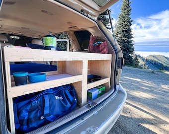 May include: The back of a white minivan with the tailgate open, revealing a custom-built wooden shelf system. The shelves are filled with camping gear, including a blue duffel bag, a green propane tank, and blue cups. The van is parked in a scenic mountain setting with a view of a cloudy sky.