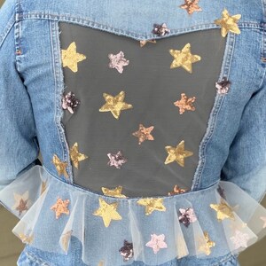 Pode incluir: Uma jaqueta de ganga azul com um painel de malha transparente nas costas. O painel de malha é decorado com lantejoulas em forma de estrela douradas, cor-de-rosa e castanhas. A jaqueta tem uma bainha com babados com uma sobreposição de tule que também é decorada com lantejoulas em forma de estrela.