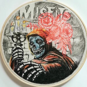 May include: Embroidered hoop art featuring a skeletal figure in a hooded robe, holding a candelabra. The figure has a blue face and a toothy grin. The background is a gray brick wall with the word "MISFIT" written in black. A cluster of red flowers is embroidered above the figure's head.