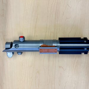 Puede incluir: Un sable de luz plateado con un mango negro y un bot&oacute;n rojo en la parte superior. El sable de luz tiene un detalle marr&oacute;n y plateado en el medio.