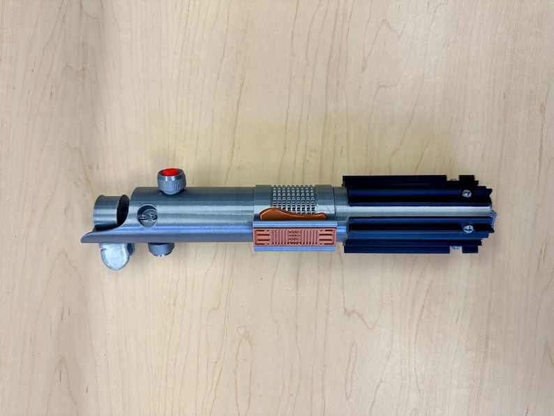 Puede incluir: Un sable de luz plateado con un mango negro y un bot&oacute;n rojo en la parte superior. El sable de luz tiene un detalle marr&oacute;n y plateado en el medio.