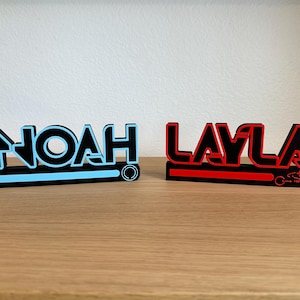 Può includere: Due insegne con nomi personalizzati, "Noah" in blu e nero e "Layla" in rosso e nero. Ogni insegna è un design stampato in 3D con un carattere unico e un elemento decorativo alla base, adatto per l'arredamento della stanza.