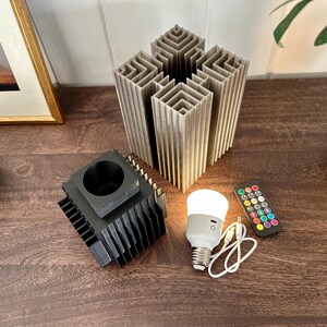 May include: A black and grey 3D printed lamp base with a white smart bulb and a remote control. The lamp base is made of multiple layers of stacked squares.