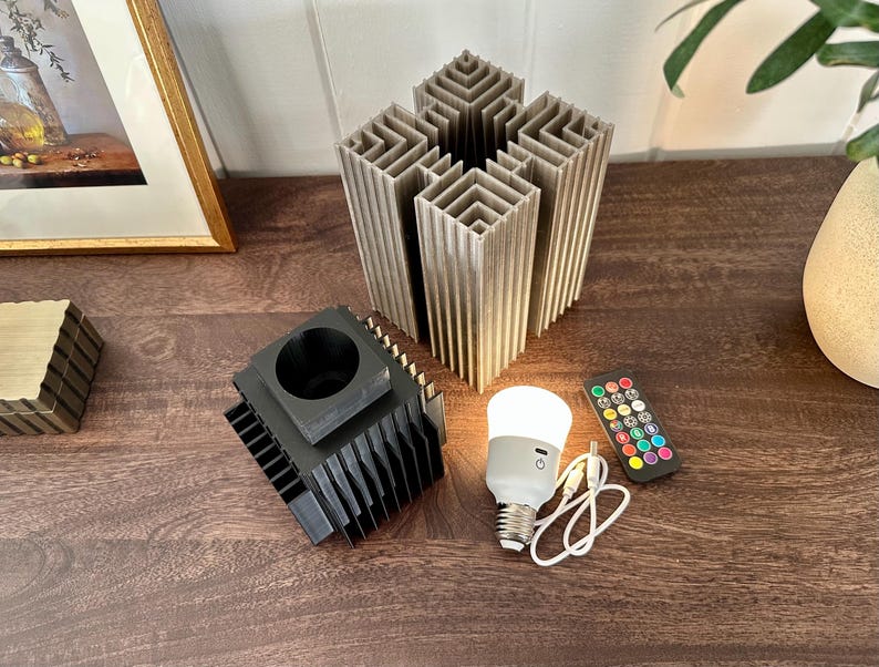 May include: A black and grey 3D printed lamp base with a white smart bulb and a remote control. The lamp base is made of multiple layers of stacked squares.