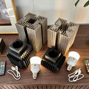 May include: Two black light bulb holders with a geometric design, each with a white smart bulb. A remote control with coloured buttons is on the table next to the light bulb holders.