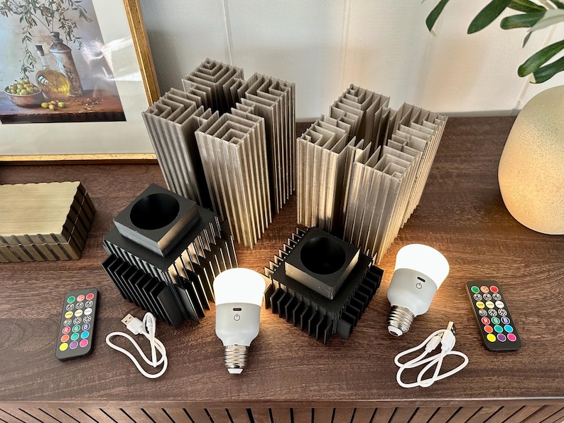 May include: Two black light bulb holders with a geometric design, each with a white smart bulb. A remote control with coloured buttons is on the table next to the light bulb holders.