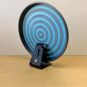 May include: A round, blue and gray decorative plate with concentric circles, supported by a black metal stand. The plate has a dark gray rim and a light blue center. The stand is labeled "amazonbasics".