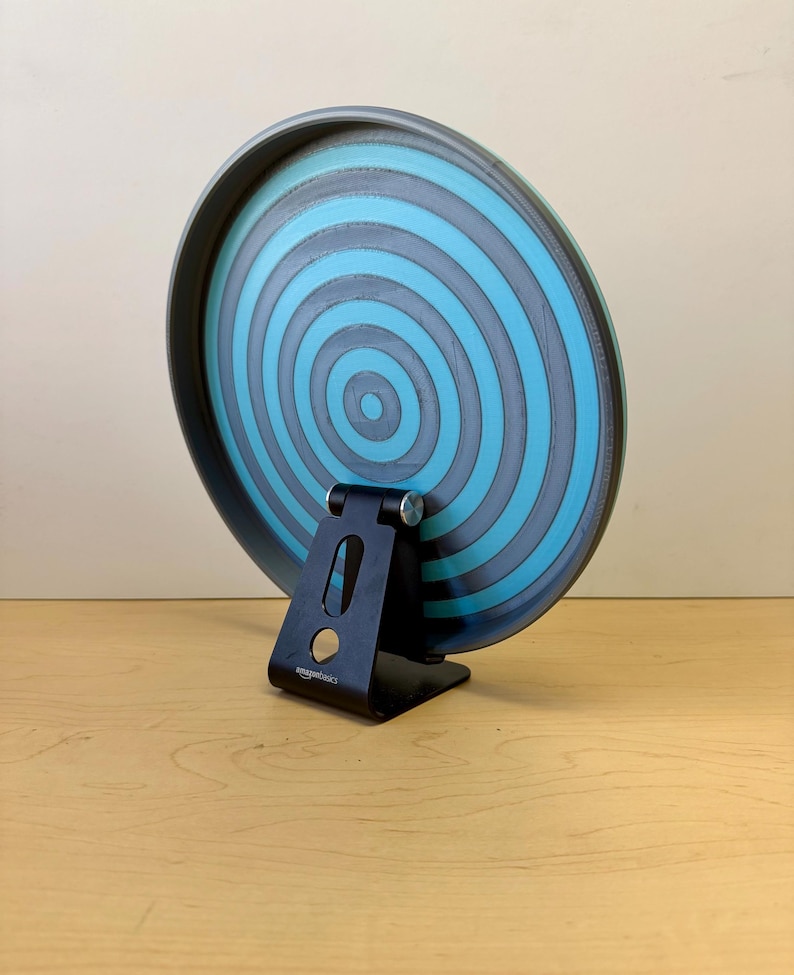 May include: A round, blue and gray decorative plate with concentric circles, supported by a black metal stand. The plate has a dark gray rim and a light blue center. The stand is labeled "amazonbasics".