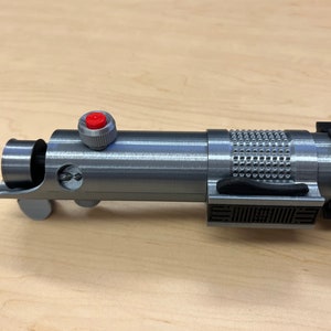 Puede incluir: Una r&eacute;plica de sable de luz plateado con un mango negro y un bot&oacute;n rojo. El sable de luz tiene un agarre texturizado y un detalle negro en el mango.