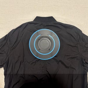 May include: Black bomber jacket with a circular, futuristic design attached to the back. The design features a black center with a blue outer ring and a series of concentric circles. The jacket is made of a smooth, shiny material.