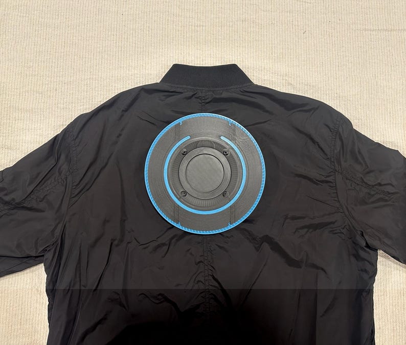 May include: Black bomber jacket with a circular, futuristic design attached to the back. The design features a black center with a blue outer ring and a series of concentric circles. The jacket is made of a smooth, shiny material.
