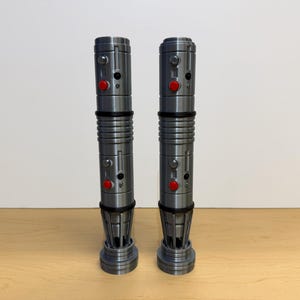May include: Two metallic gray lightsabers with red buttons and black accents are displayed on a wooden surface. The lightsabers have a cylindrical design with detailed ridges and a flared base. The background is a plain white wall.