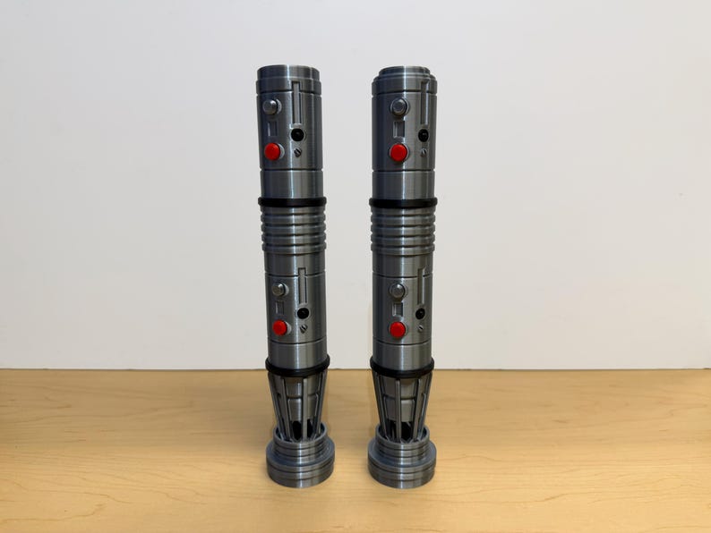 May include: Two metallic gray lightsabers with red buttons and black accents are displayed on a wooden surface. The lightsabers have a cylindrical design with detailed ridges and a flared base. The background is a plain white wall.