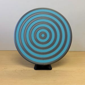May include: A round, gray and teal object with a concentric circle design. The object is mounted on a small black stand and sits on a light wood surface. The design features alternating bands of gray and teal.