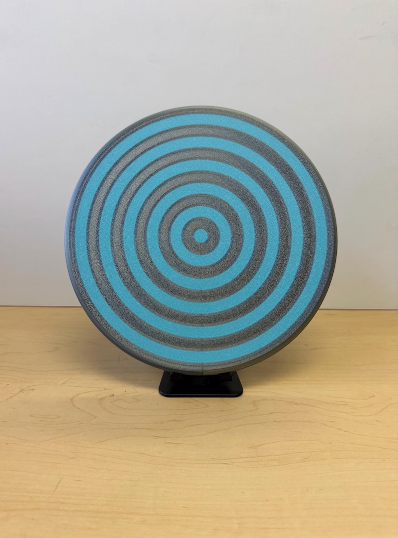 May include: A round, gray and teal object with a concentric circle design. The object is mounted on a small black stand and sits on a light wood surface. The design features alternating bands of gray and teal.