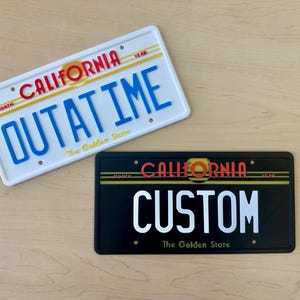 May include: Two California license plates. The top plate is white with black and red text that reads "CALIFORNIA OUTATIME The Golden State". The bottom plate is black with white and red text that reads "CALIFORNIA CUSTOM The Golden State".