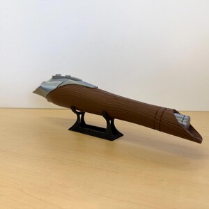 May include: A brown, cylindrical object with a silver accent and a black stand. The object has a textured surface and a tapered end. It is displayed on a light brown surface against a white background.