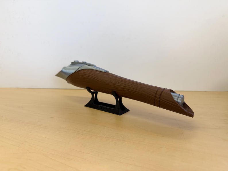 May include: A brown, cylindrical object with a silver accent and a black stand. The object has a textured surface and a tapered end. It is displayed on a light brown surface against a white background.