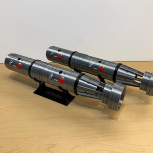 May include: Two silver and black lightsabers with red buttons are displayed on a black stand. The lightsabers have a detailed design and are resting on a light brown wooden surface.
