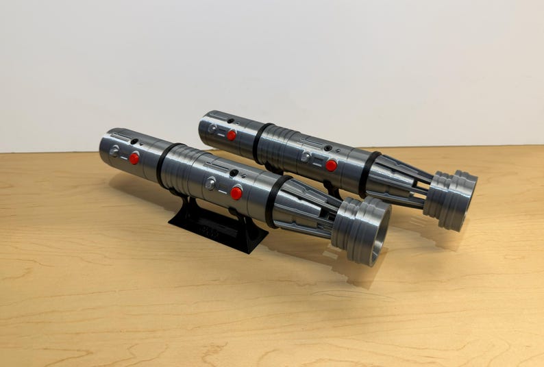 May include: Two silver and black lightsabers with red buttons are displayed on a black stand. The lightsabers have a detailed design and are resting on a light brown wooden surface.