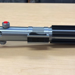 Puede incluir: Un accesorio de sable de luz plateado y negro con un bot&oacute;n rojo en el mango. El sable de luz tiene un agarre texturizado y una hoja negra.