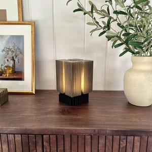 May include: A small, grey, cube-shaped lamp with a black base sits on a dark brown wooden table. The lamp has a geometric design and emits a warm glow. A vase with green branches and a framed picture are in the background.