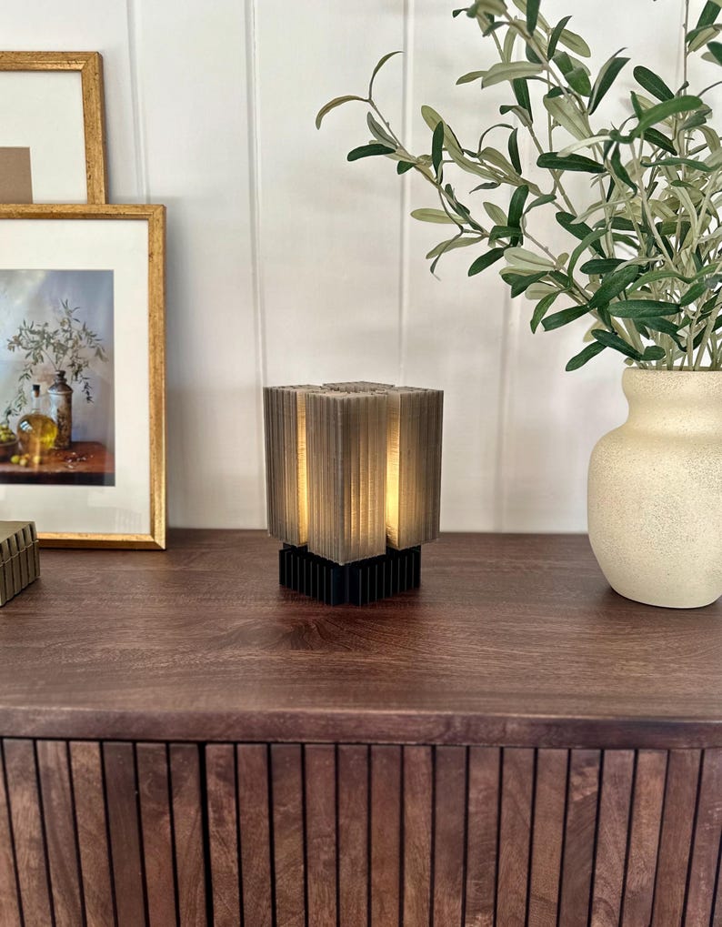 May include: A small, grey, cube-shaped lamp with a black base sits on a dark brown wooden table. The lamp has a geometric design and emits a warm glow. A vase with green branches and a framed picture are in the background.