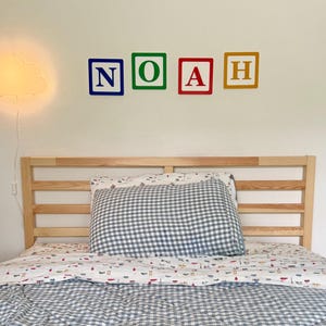 May include: A bed with a light wood headboard and a blue and white gingham patterned pillow and comforter. The word "NOAH" is spelled out in colorful block letters on the wall above the bed. A cloud-shaped lamp is on the left.