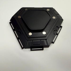 May include: A black, hexagonal device with a central rectangular panel and several circular silver accents. The device has a geometric design with multiple rectangular cutouts and a handle-like projection. The object is set against a white background.