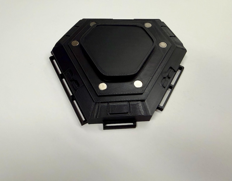 May include: A black, hexagonal device with a central rectangular panel and several circular silver accents. The device has a geometric design with multiple rectangular cutouts and a handle-like projection. The object is set against a white background.