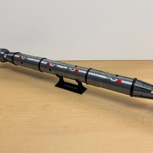 May include: A gray lightsaber prop with red buttons and black accents, resting on a black stand. The prop is cylindrical with detailed ridges and a complex end design. It is displayed on a light brown wooden surface.