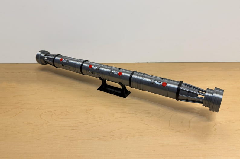 May include: A gray lightsaber prop with red buttons and black accents, resting on a black stand. The prop is cylindrical with detailed ridges and a complex end design. It is displayed on a light brown wooden surface.