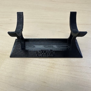May include: A black 3D-printed lightsaber stand. The stand features a rectangular base with the words "STAR WARS" embossed on the front. Two curved supports rise from the base to cradle a lightsaber. The stand is designed to hold a lightsaber.