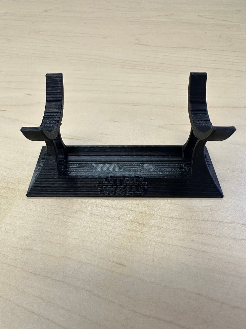 May include: A black 3D-printed lightsaber stand. The stand features a rectangular base with the words "STAR WARS" embossed on the front. Two curved supports rise from the base to cradle a lightsaber. The stand is designed to hold a lightsaber.