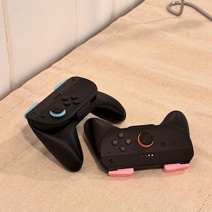 May include: Two black game controllers with light blue and pink accents. One controller has a cat ear design. The controllers are on a beige surface. The controllers have buttons and joysticks.