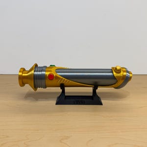 May include: A gold and gray lightsaber prop, resting on a black stand. The lightsaber features gold accents, a gray body, and red and green buttons. The prop is displayed on a light brown wooden surface.