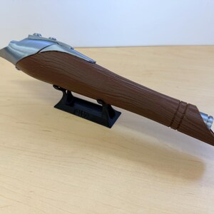 May include: A brown and silver lightsaber prop, resting on a black stand. The lightsaber has a textured brown handle and a silver blade guard. The stand has the text "STAR WARS" on it.