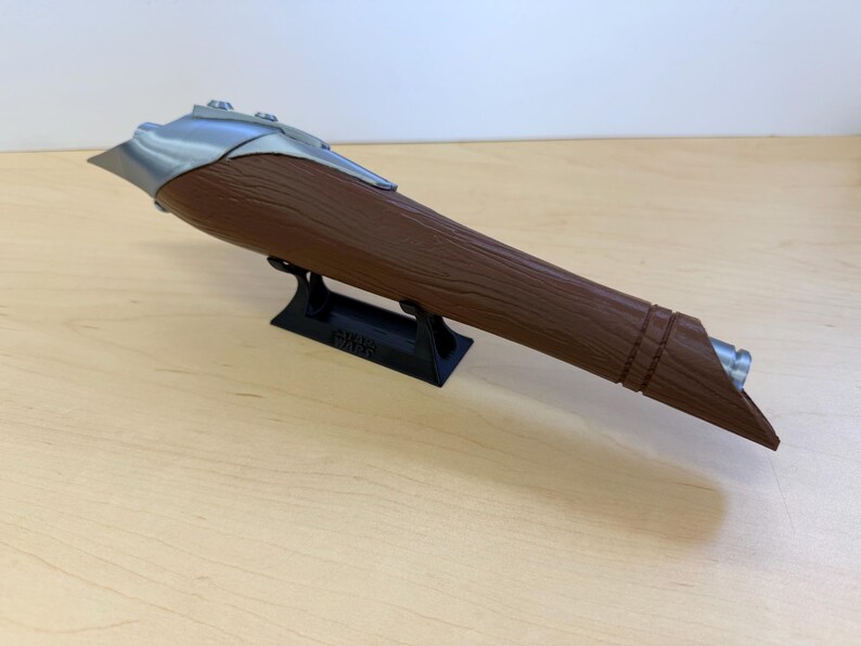 May include: A brown and silver lightsaber prop, resting on a black stand. The lightsaber has a textured brown handle and a silver blade guard. The stand has the text "STAR WARS" on it.