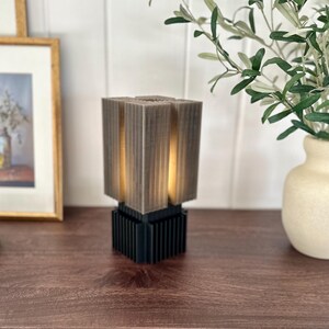 May include: A modern, geometric table lamp with a grey, textured shade and a black base. The lamp is lit and casting a warm glow.