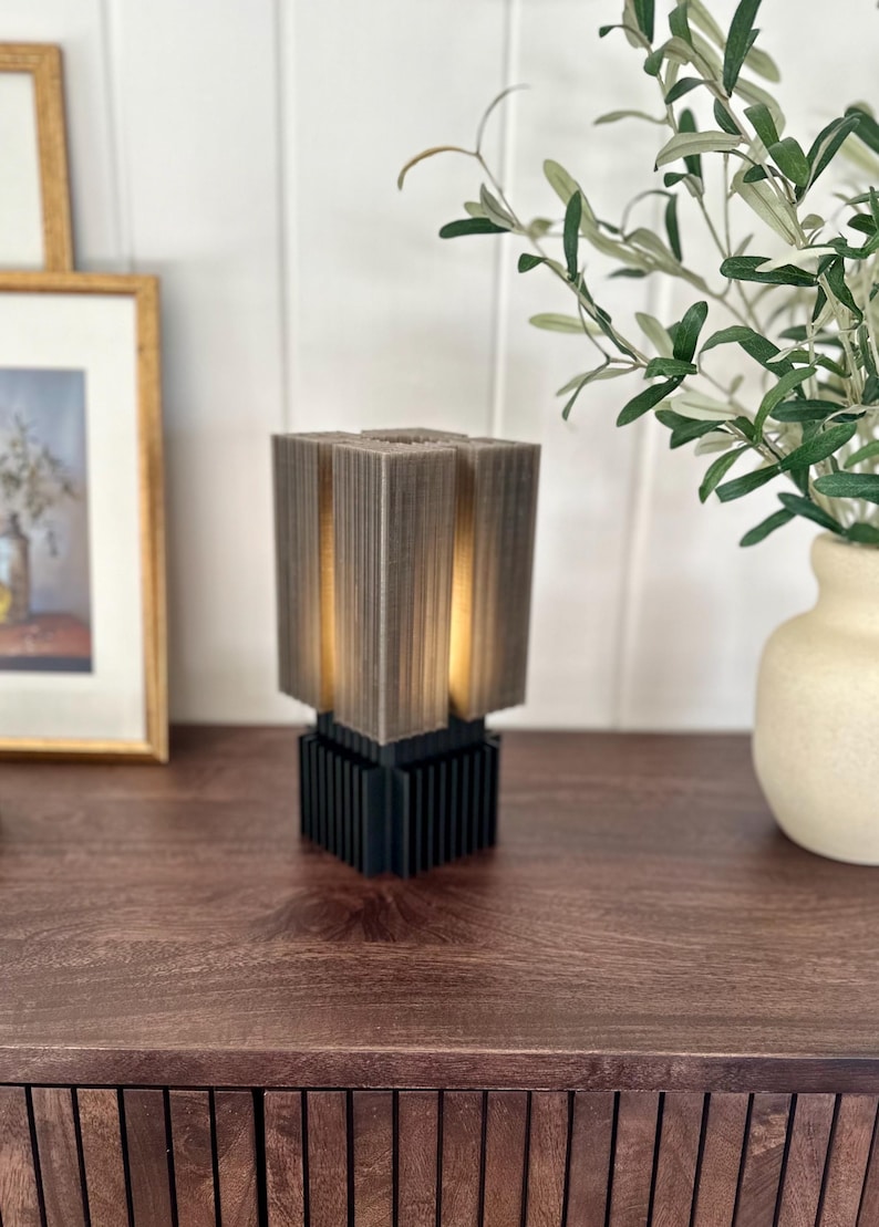 May include: A modern, geometric table lamp with a grey, textured shade and a black base. The lamp is lit and casting a warm glow.