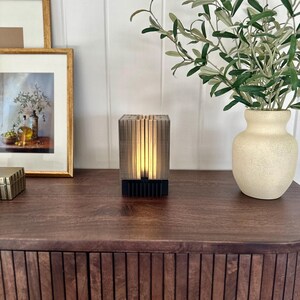 May include: A modern, wooden table lamp with a warm, ambient glow. The lamp has a rectangular base and a geometric design. The lamp is sitting on a dark wood table with a vase of greenery in the background.