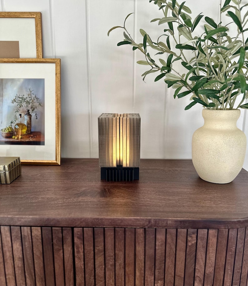 May include: A modern, wooden table lamp with a warm, ambient glow. The lamp has a rectangular base and a geometric design. The lamp is sitting on a dark wood table with a vase of greenery in the background.