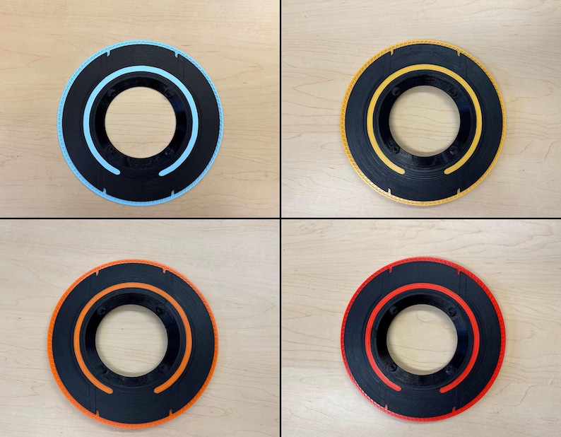 May include: Four circular, black objects with colored inner and outer rings. The rings are blue, yellow, orange, and red. Each object has a central hole and a textured outer edge. The objects are displayed on a light wood surface.