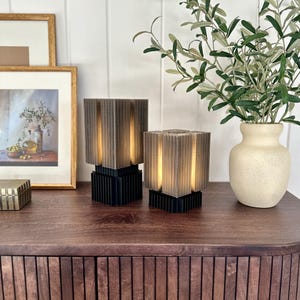 May include: Two brown and black table lamps with a ribbed design on a wooden table with a vase of green leaves.