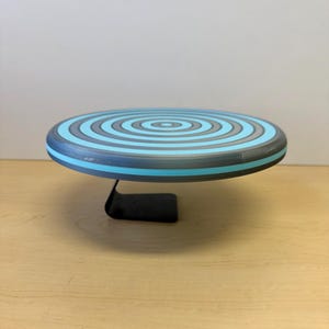 May include: A round, decorative cake stand with a blue, gray, and black spiral design. The stand is supported by a black metal base. The stand is approximately 10 inches in diameter.