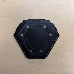 May include: A black, hexagonal object with a recessed center and several small, silver-colored circular magnets. The object has a geometric design and is set against a light-colored wooden surface.