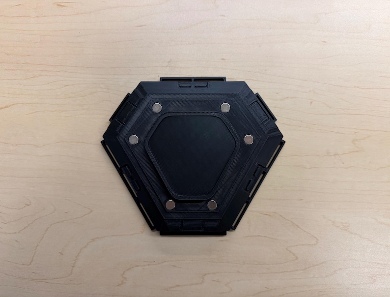 May include: A black, hexagonal object with a recessed center and several small, silver-colored circular magnets. The object has a geometric design and is set against a light-colored wooden surface.
