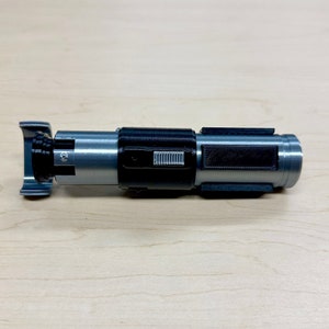 May include: A silver and black lightsaber prop with a black switch and a textured grip.