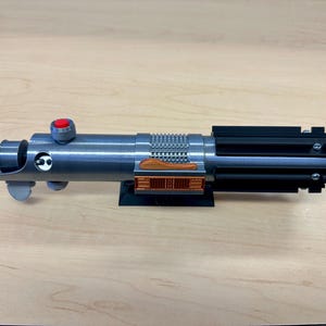 Puede incluir: Una r&eacute;plica de sable l&aacute;ser plateado con un mango negro y un detalle de color cobre. El sable l&aacute;ser est&aacute; montado en un soporte negro.